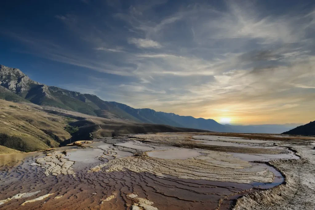 Badab-e Surt Sunset over mountain range with tiered sediment formations in the foreground and a cloudy sky above.