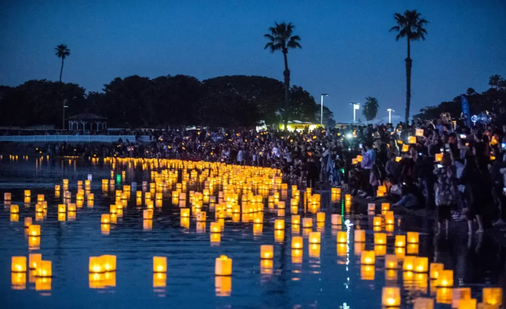 A large crowd gathers by a water's edge during twilight at the Baton Rouge, LA Water Lantern Festival, releasing numerous glowing paper lanterns onto the water. Palm trees are visible in the background, and the flickering lanterns create a warm, serene atmosphere against the dusky blue sky.