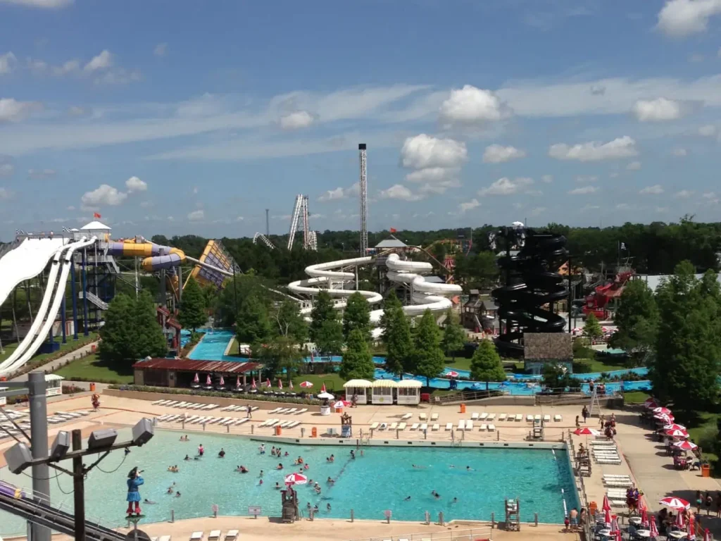 Aerial view of Blue Bayou Water Park, showcasing numerous pools, exhilarating water slides, and meandering lazy rivers. People are swimming and sunbathing. The park is bustling with activity and surrounded by lush green trees under a partly cloudy sky.