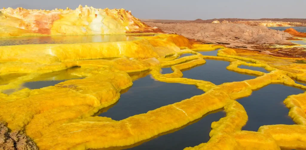 Bright yellow sulfur formations surrounding turquoise pools of water amid a rocky, barren landscape under a clear sky in the Danakil Depression, Ethiopia. The vibrant colors contrast starkly with the rugged terrain, creating a surreal, otherworldly scene that captivates all who visit.
