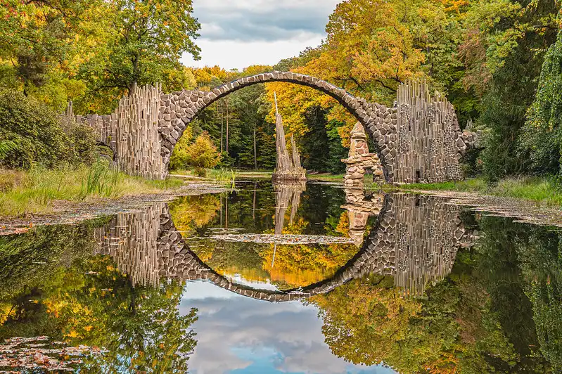 An arched stone bridge, known as Die Rakotzbrück in Germany, spans a tranquil pond surrounded by lush autumn foliage. The bridge and nearby monument perfectly reflect in the calm water, creating a circular illusion against a backdrop of vibrant trees and a cloudy sky.