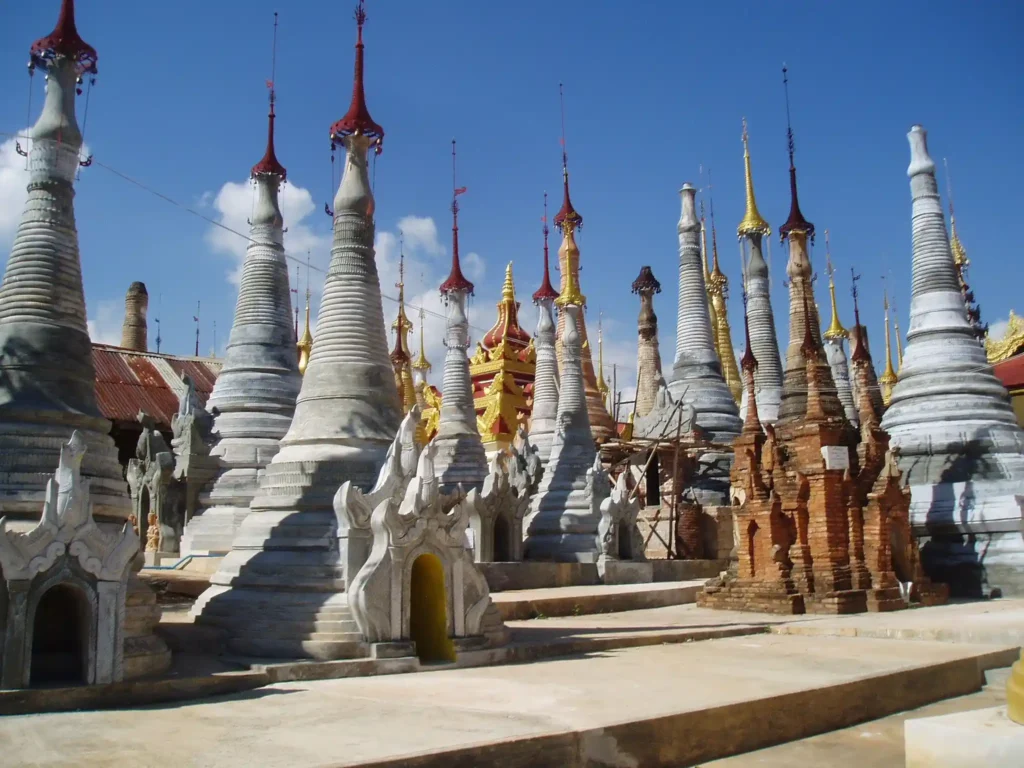 A cluster of ancient, intricately designed stupas under a clear blue sky in Nyaung Ohak, Myanmar. Some are made of stone and others red brick. Each stupa is topped with a slender, spire-like ornament. The area appears serene and historical, exuding an air of cultural and architectural significance.
