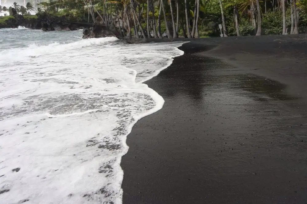 A serene black sand beach with gentle ocean waves washing onto the shore, Oneuli Beach features lush greenery and tall palm trees lining the background, creating a tropical atmosphere under a cloudy sky.