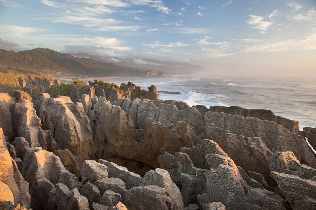 Rock formations resembling stacked pancakes, known as Pancake Rocks, are seen in the foreground. A coastline extends along the left side, with waves crashing against the shore. Green hills and misty mountains stretch into the distance under a partly cloudy sky.
