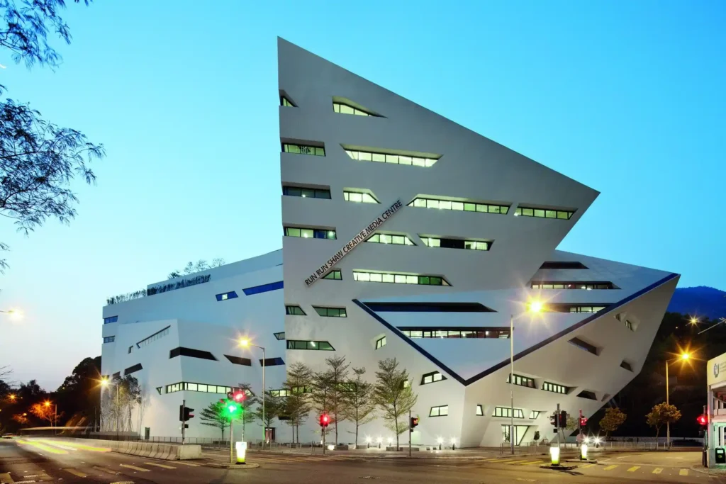 The Shaw Center for the Arts is a modern angular building with sharp edges and a white façade. It features large, irregularly-shaped windows and is illuminated by streetlights in the evening. There are trees and traffic lights in front of the building, set against a clear sky.