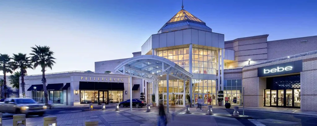 The Mall of Louisiana, a modern shopping destination, features a striking glass pyramid roof and entrance canopy surrounded by storefronts such as Pottery Barn and Bebe. Palm trees line the walkway, where several people stroll near parked cars at dusk against a backdrop of a blue sky.