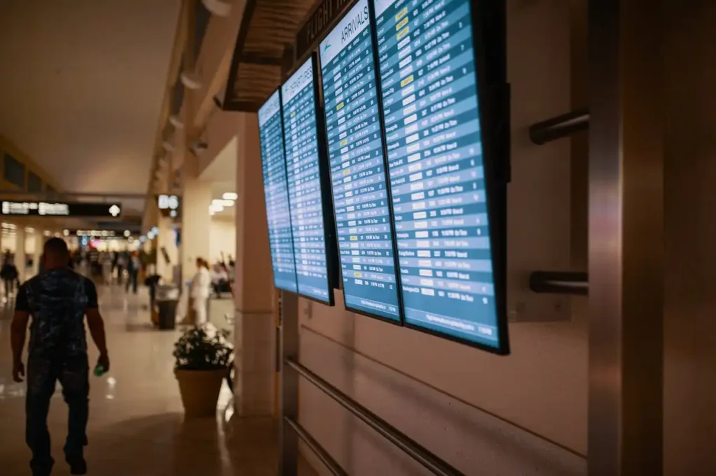 Digital flight information displays at an airport show arrival and departure times. Passengers can be seen walking in the background. The terminal's clean, well-lit space features beige walls and a few potted plants. The atmosphere appears busy and organized.