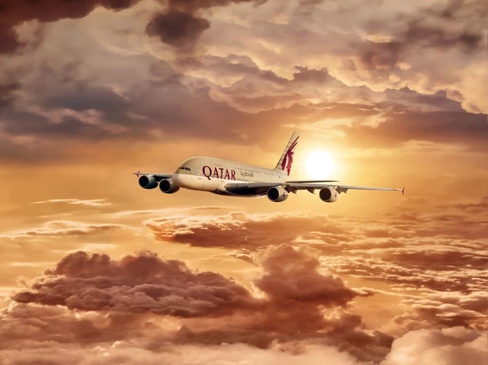 A Qatar Airlines plane flies mid-air against a dramatic sunset backdrop with golden and reddish clouds. The plane is prominently seen with the airline's logo on its body and the sun partially visible near the tail.