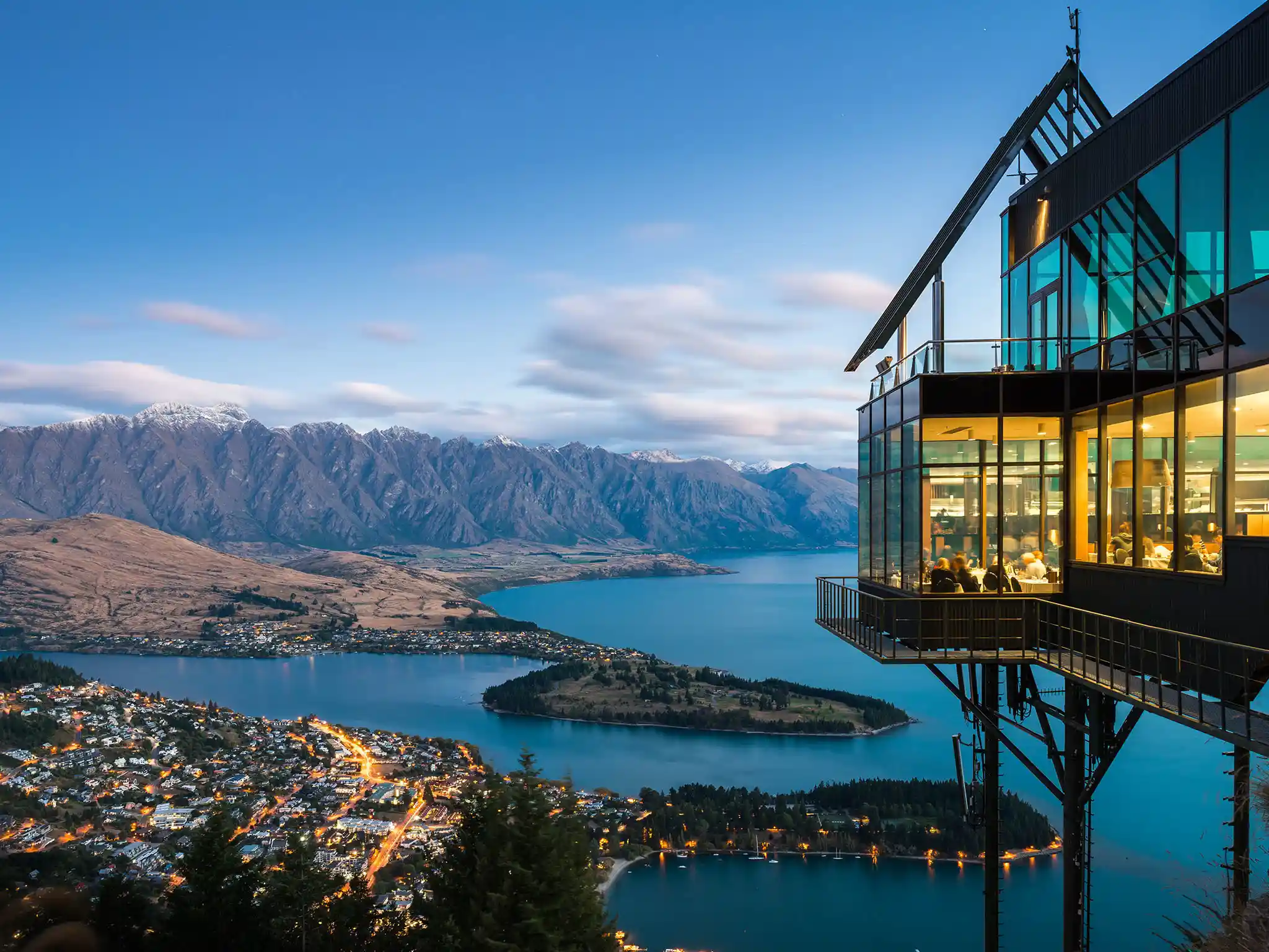 Evening shot overlooking the lakeside town of Queenstown, featuring a mountainside building with large glass windows. The landscape includes a sprawling lake, a small island, illuminated streets, and mountains in the background under a partly cloudy New Zealand sky.