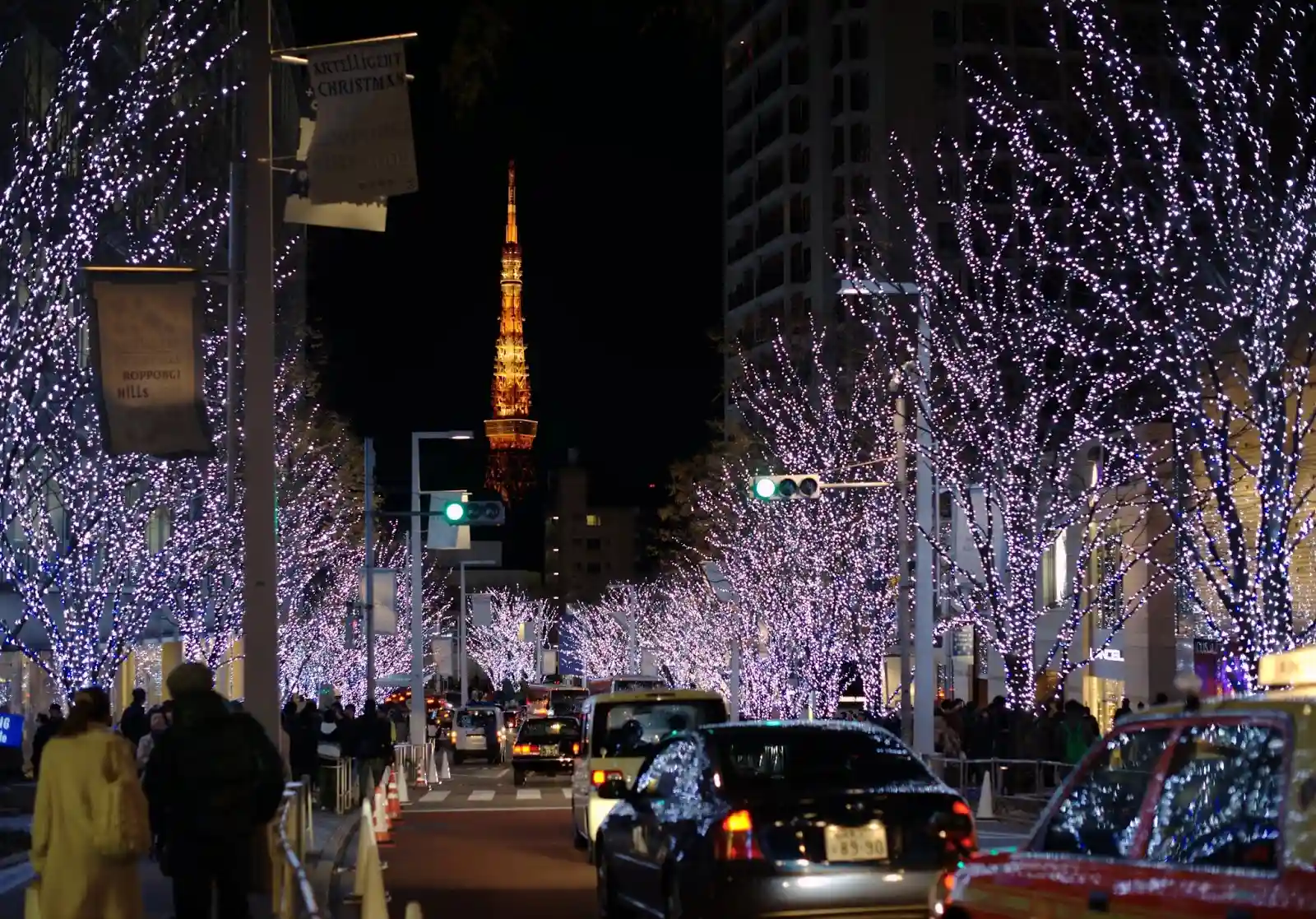 A bustling city street in Tokyo adorned with trees wrapped in twinkling white lights on both sides. The street is crowded with people and cars. In the background, a tall, illuminated tower glows golden against the night sky.