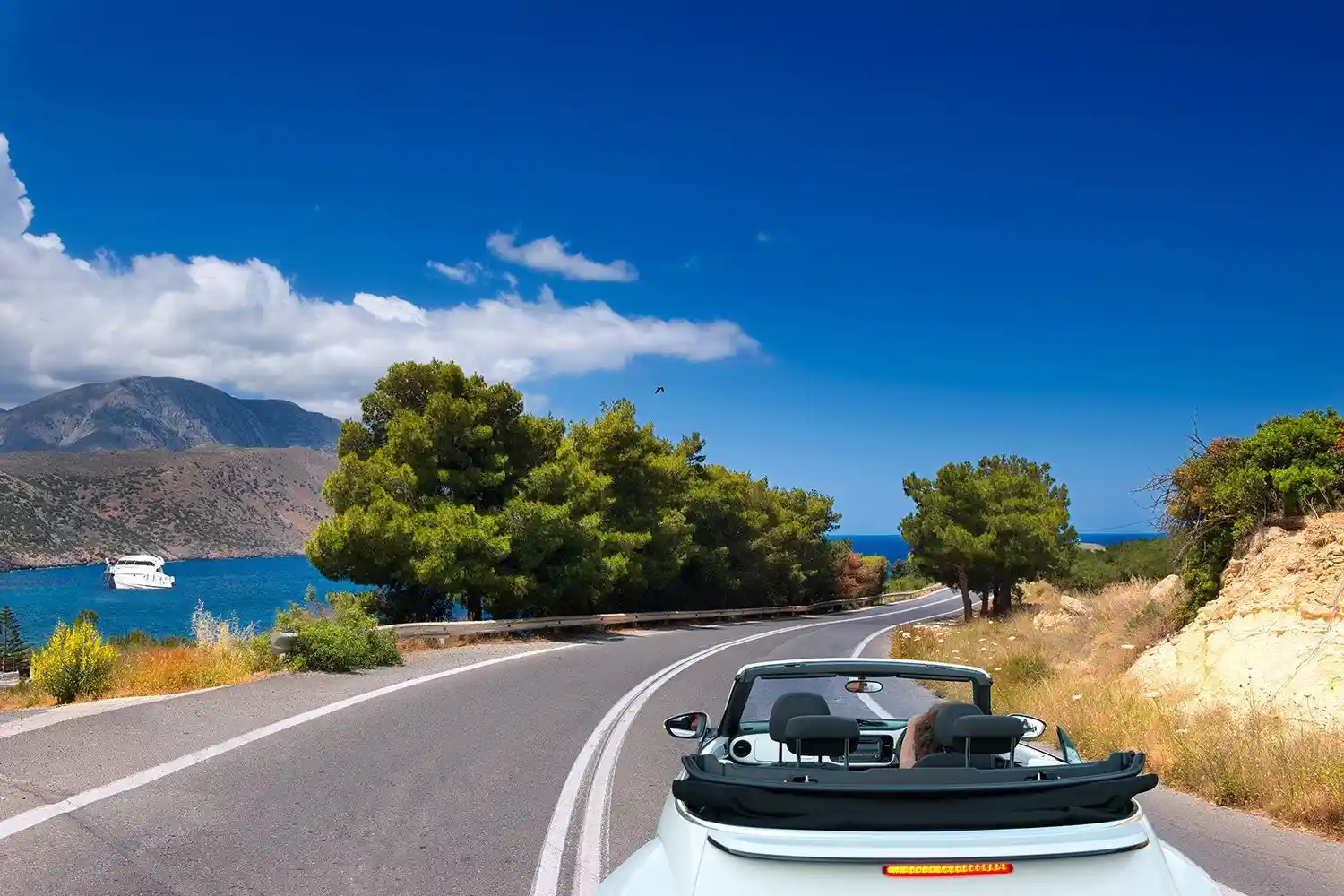 A white convertible drives along a scenic coastal road lined with trees. The road curves with a view of the blue sea and a distant boat. The sky is clear with a few white clouds in the background.